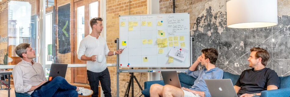 three men sitting while using laptops and watching man beside whiteboard